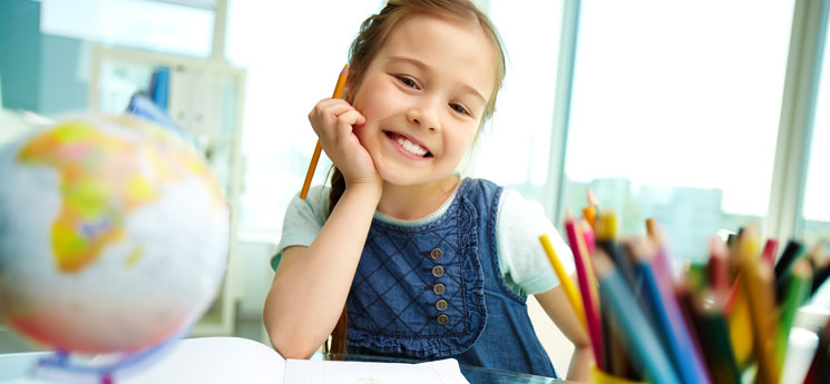 young girl smiling while holding colored pencil and working at desk in fancy classroom