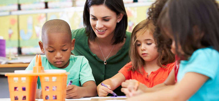 young teacher watching over three preschoolers working at craft table in classroom